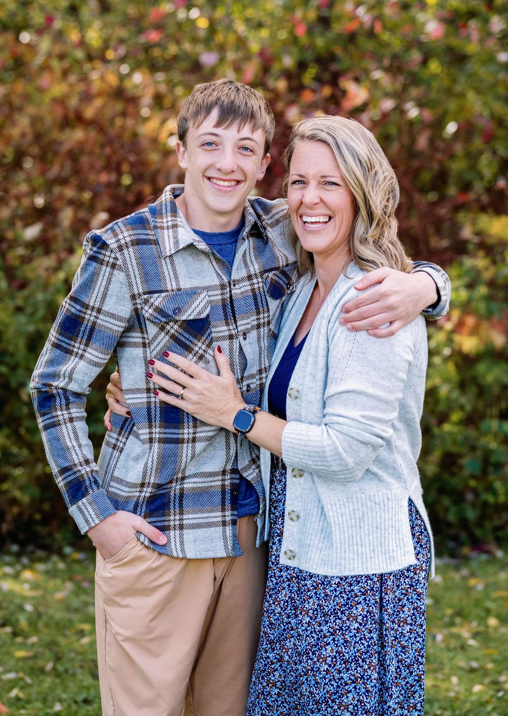 A smiling young man and woman pose together outdoors in fall, wearing casual layered outfits with colorful foliage behind them.