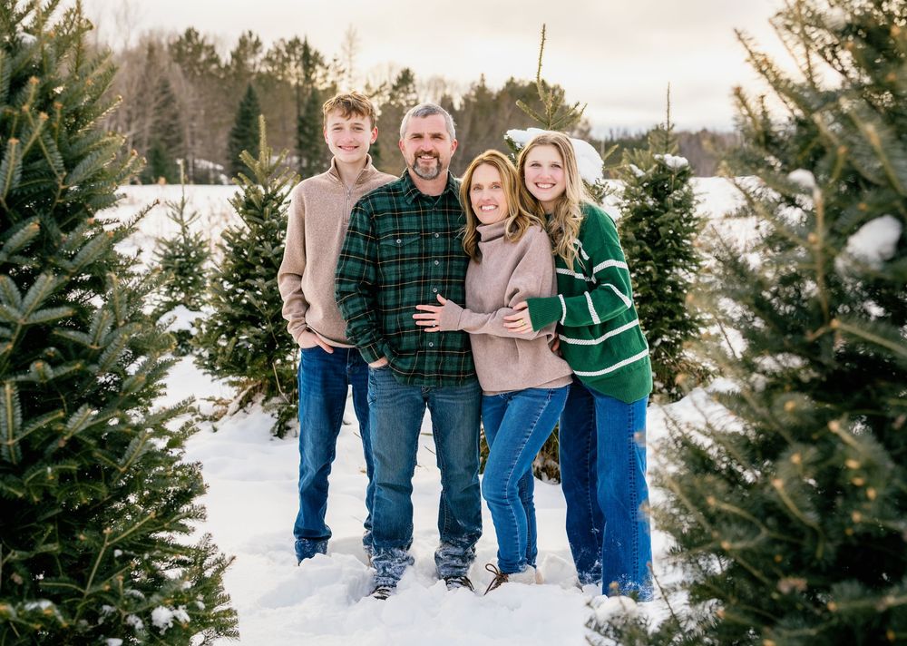 Family of four smiling together at a snowy Christmas tree farm, wearing cozy winter outfits.