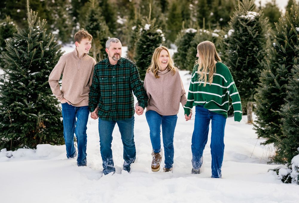 Family of four walking through snowy Christmas tree farm, smiling and holding hands in festive winter sweaters.