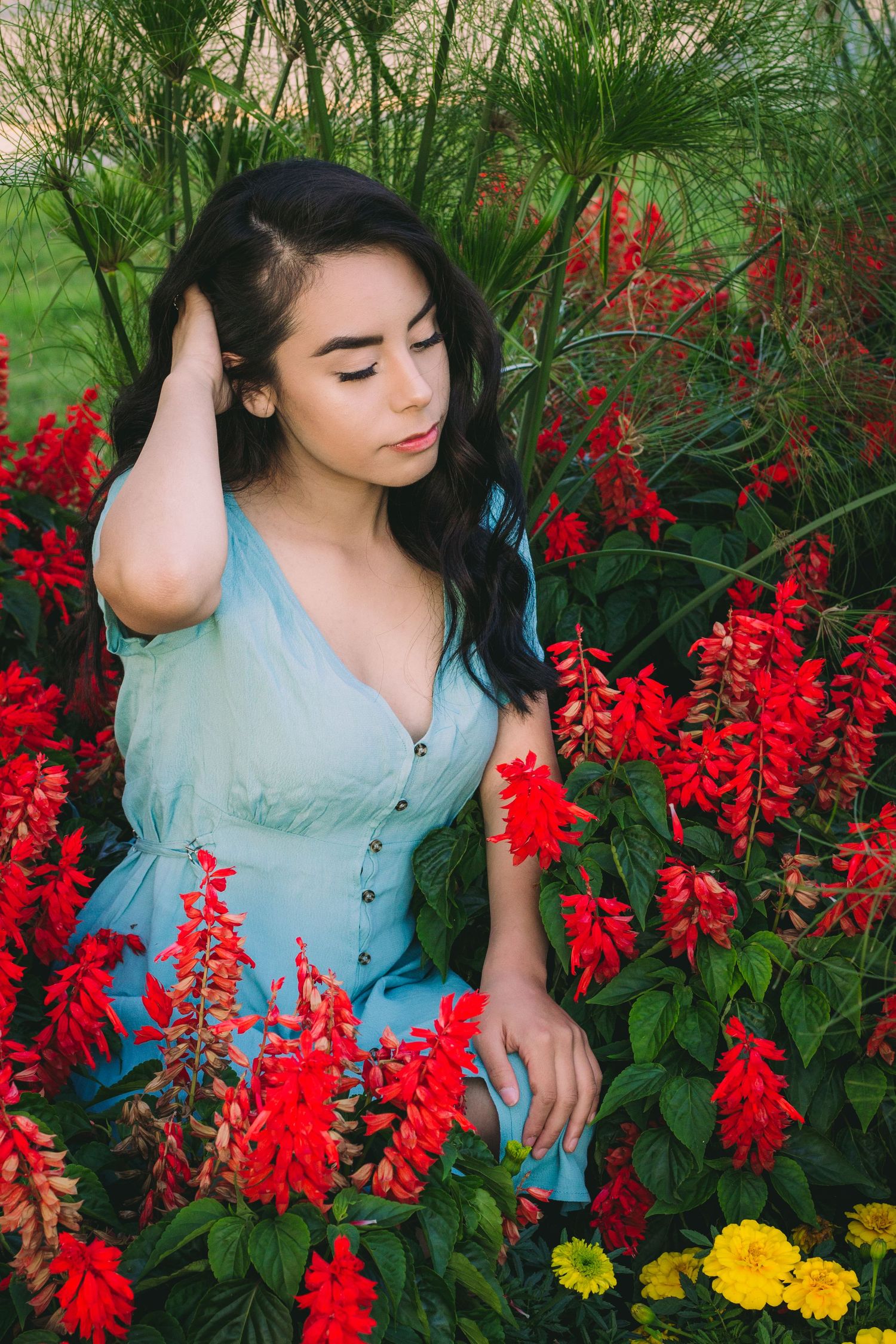Person in light blue top poses gracefully among vibrant red flowers and greenery in garden setting.
