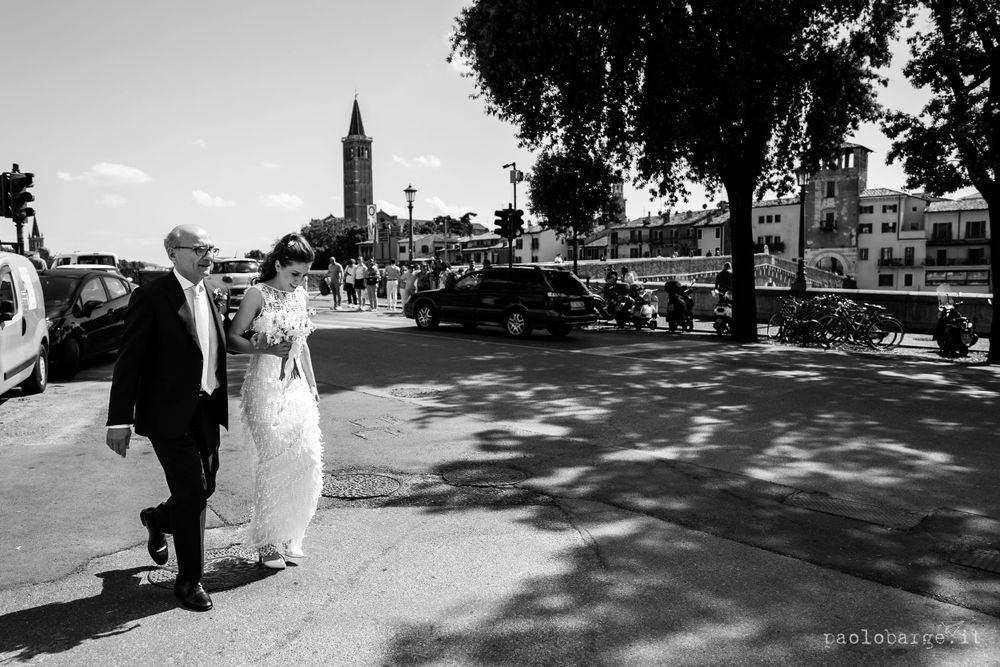 A couple walks along a sunny street with historic buildings and a church tower in the background.