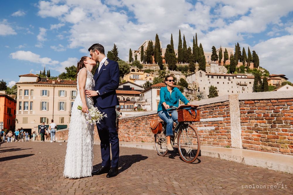 Wedding couple poses against backdrop of cypress trees and Italian hillside buildings.