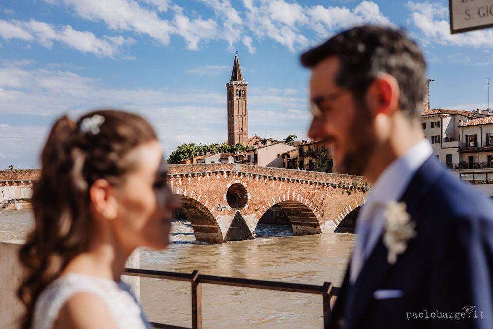 Wedding photo with historic Verona bridge and tower visible in the background on a sunny day.