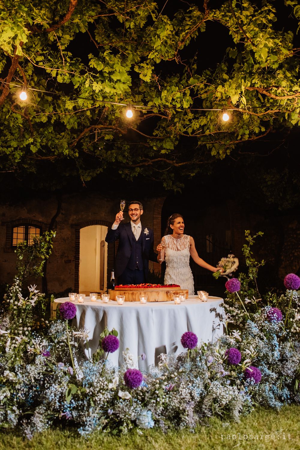 Reception table decorated with purple flowers and string lights under leafy garden canopy.