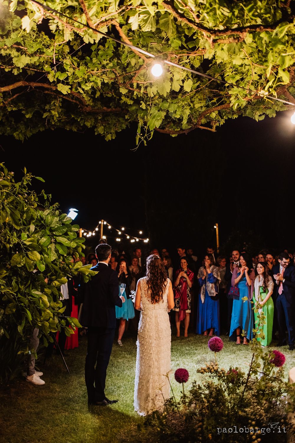 Wedding celebration under string lights with guests gathered in a garden setting.