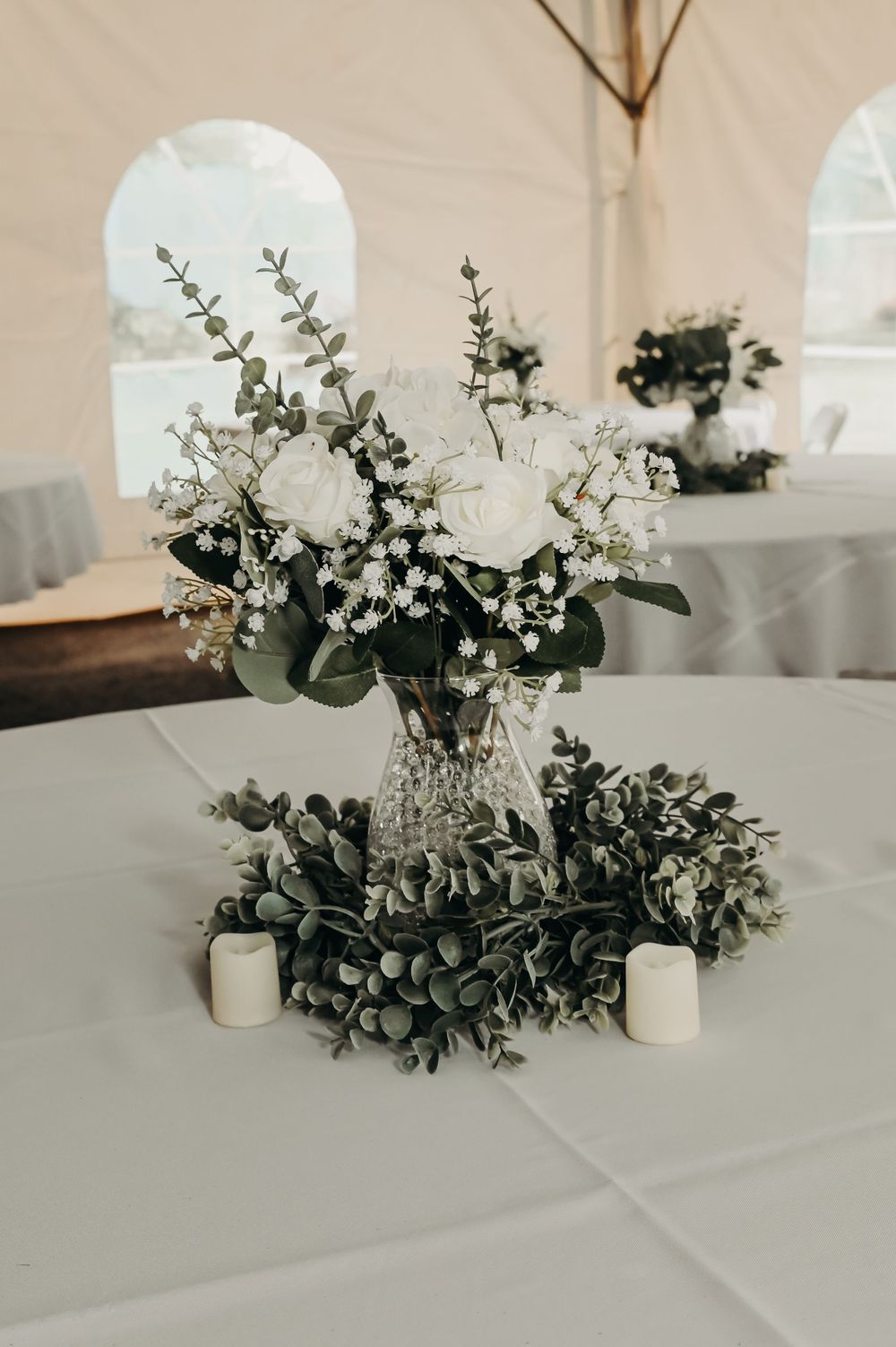 Elegant white rose and baby's breath centerpiece in crystal vase with greenery on reception table.