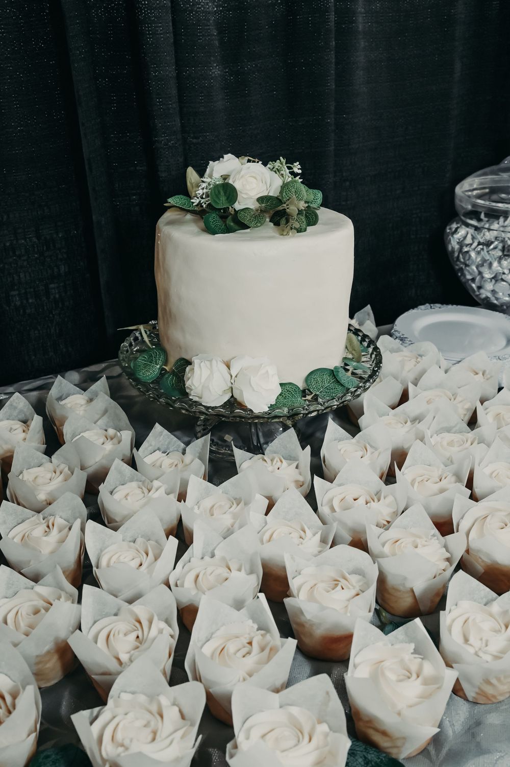 White wedding cake with rose decorations surrounded by matching cupcakes arranged on display.
