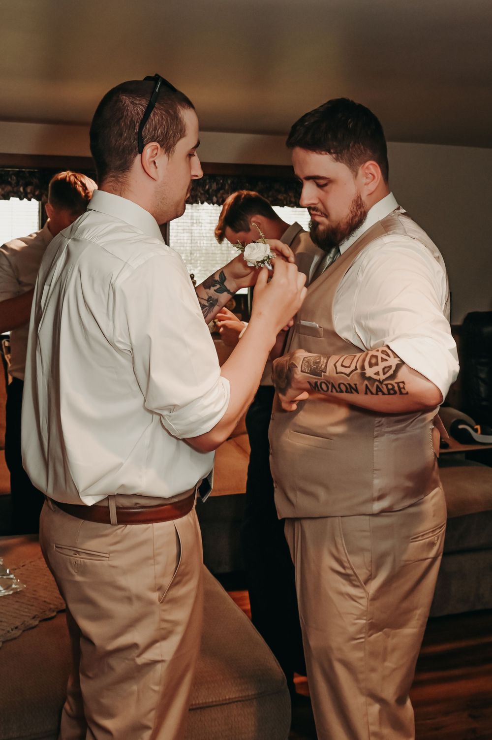 Two groomsmen in matching khaki pants and white shirts help each other with final wedding preparations.