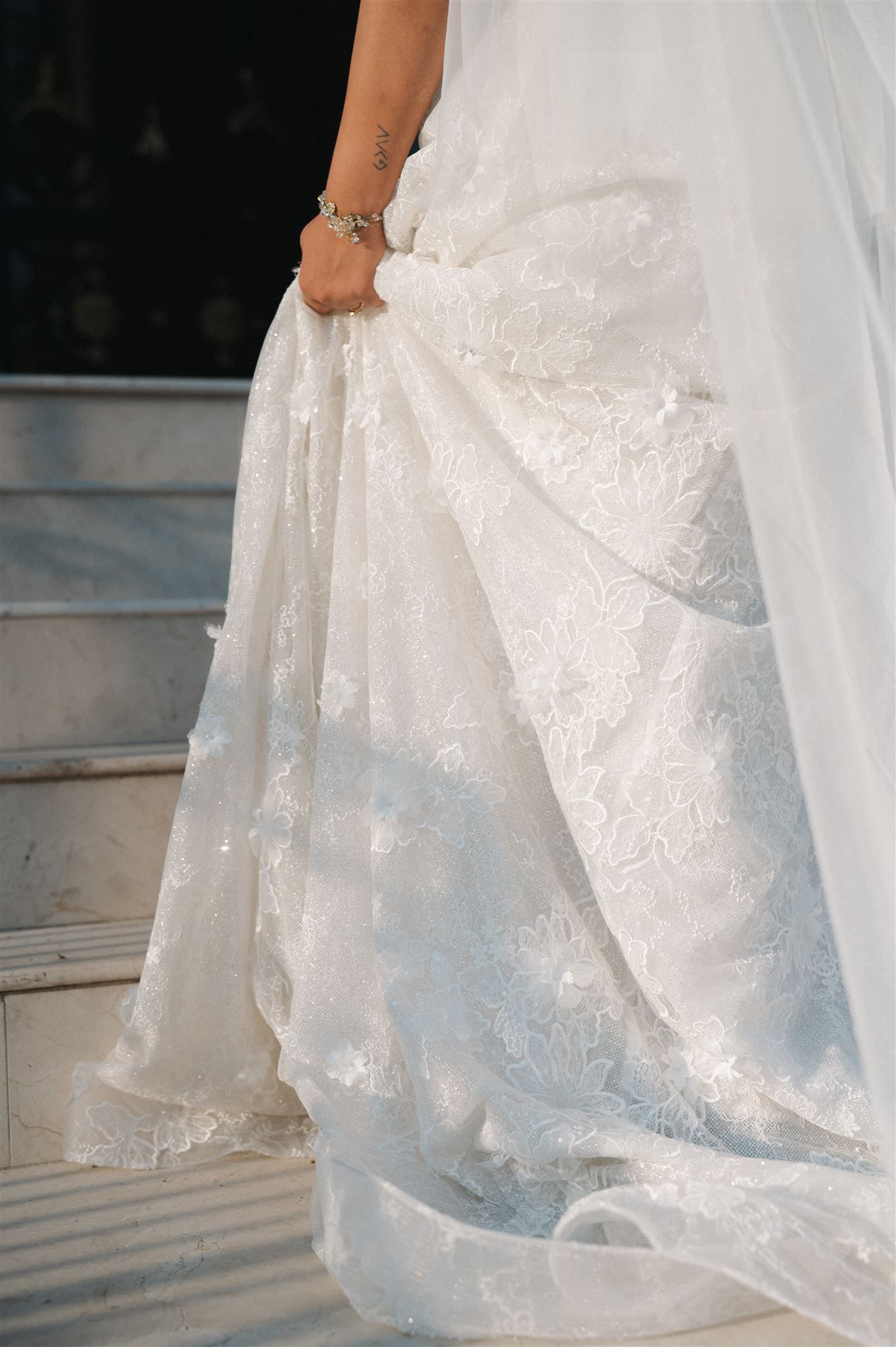 A close-up of an elegant white lace wedding dress with delicate details being gathered on marble steps.