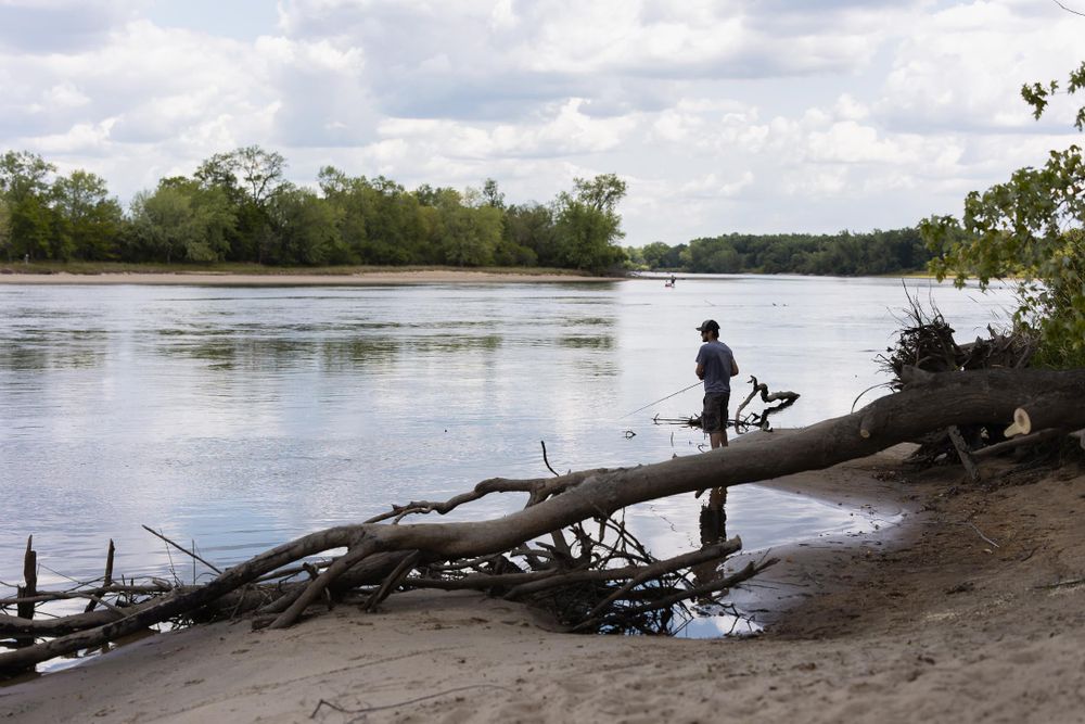 landscape photo of river with green trees and sandy shore with man fishing in distance