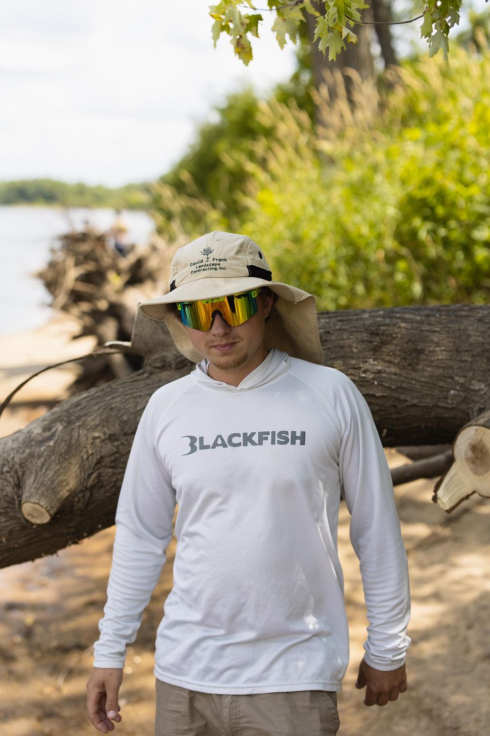 man in white shirt wearing sunglasses and fisherman's hat walking on sandy beach by river