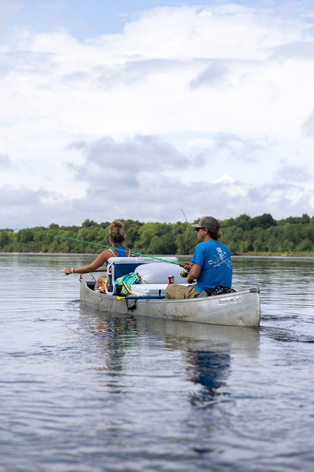 man and woman on canoe on river with man holding fishing rod and canoe packed with camping gear