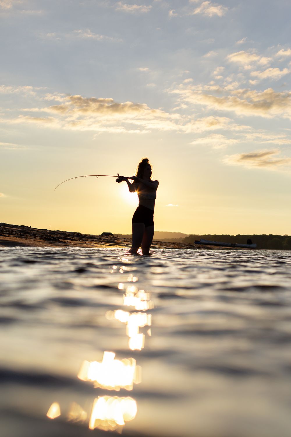 lady with hair in bun standing in water casting fishing rod with sunset behind