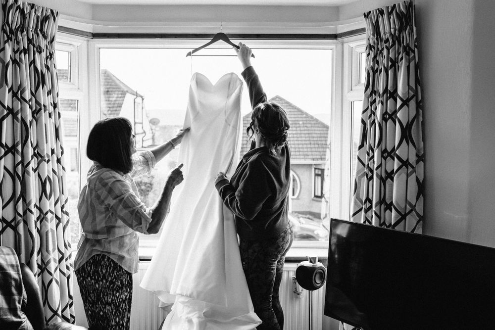 Two people examining white wedding dress hanging in window with patterned curtains.