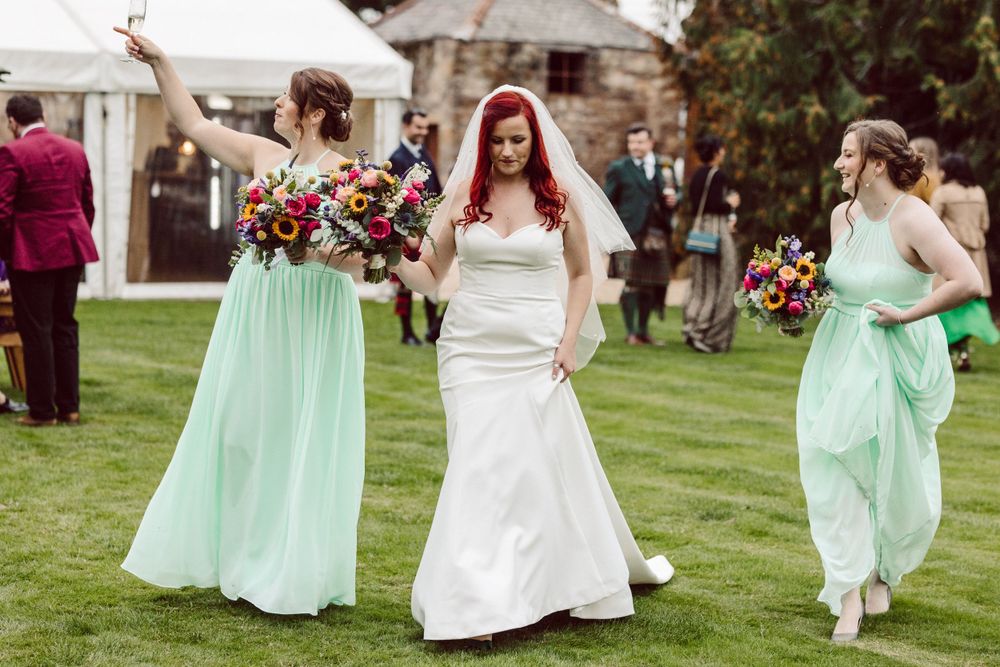 Three wedding party members in mint green dresses celebrate joyfully on a lawn.