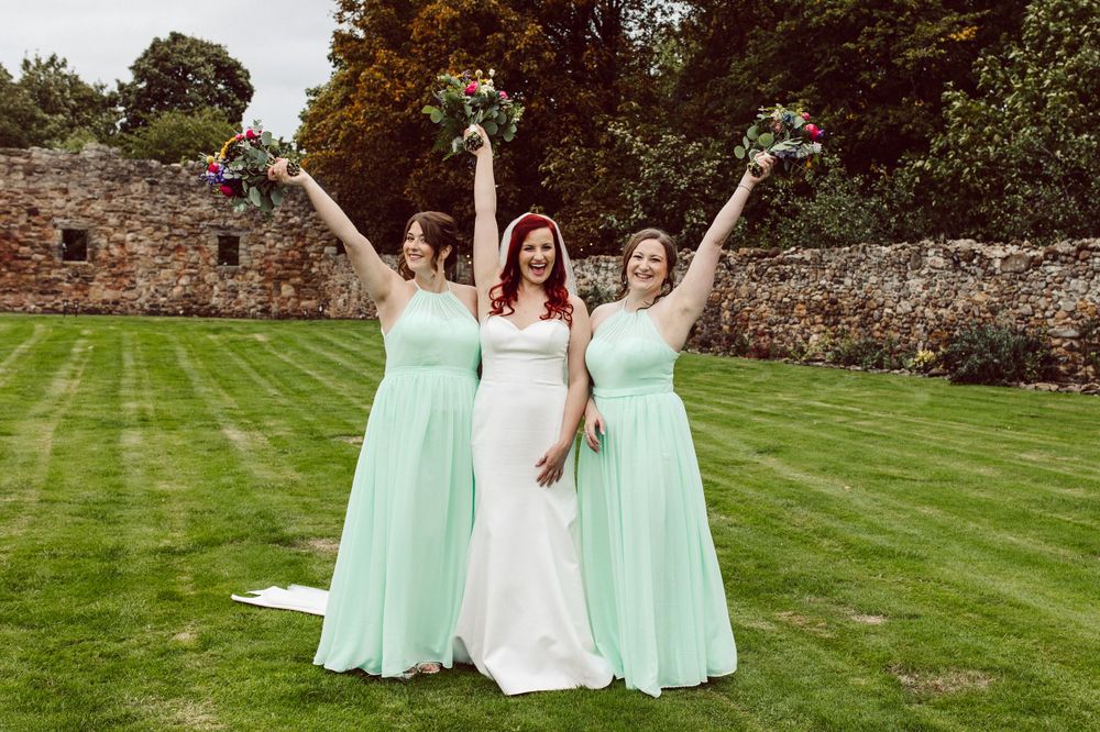 Three women in wedding attire raise their bouquets triumphantly on a green lawn.