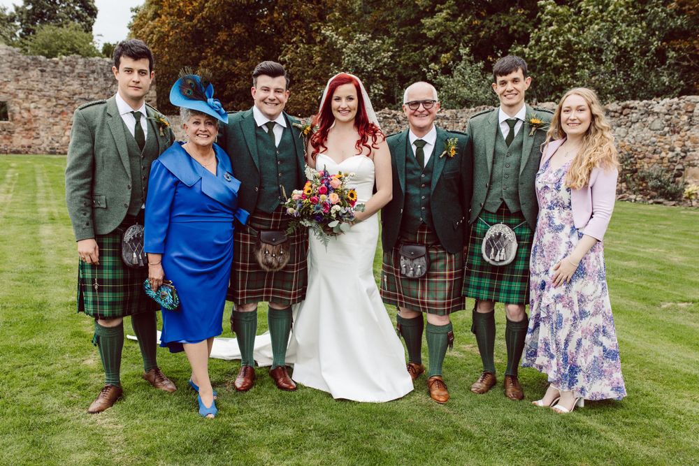 Wedding party poses on green lawn wearing traditional Scottish kilts and formal attire.