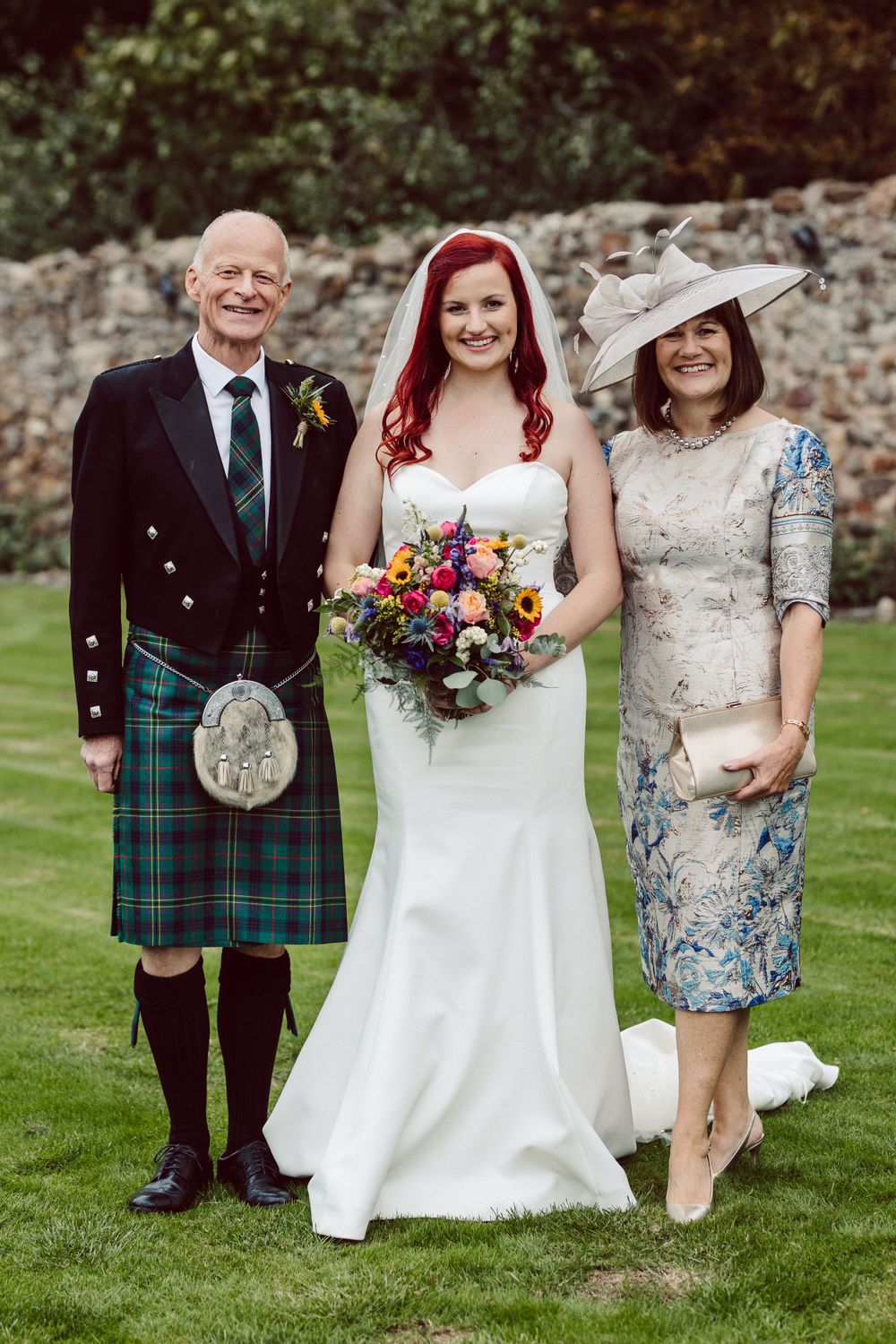 Wedding party members in Scottish formal wear and kilts pose on manicured lawn.