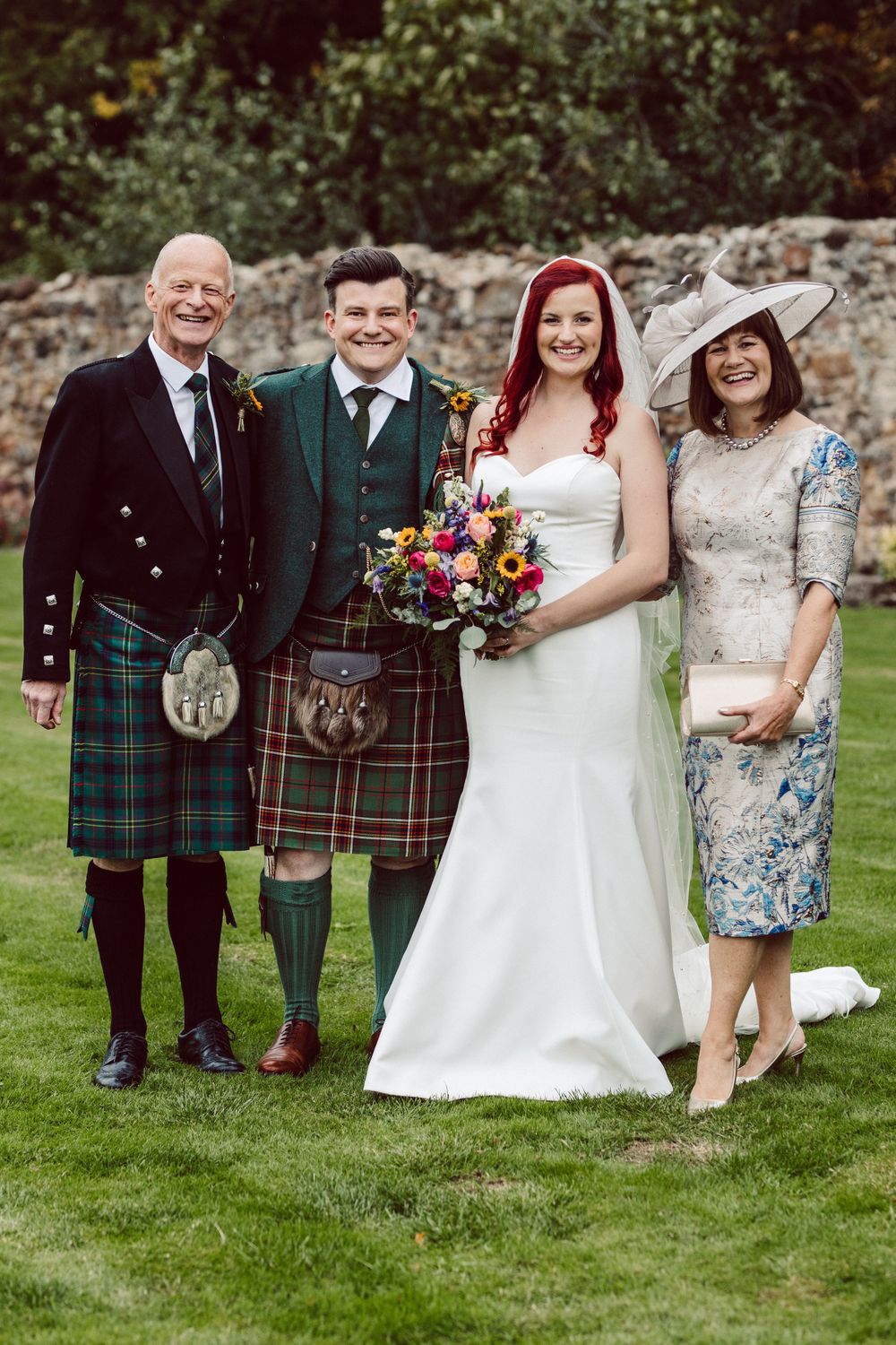 Wedding party members in traditional Scottish attire gather for formal photos outdoors.