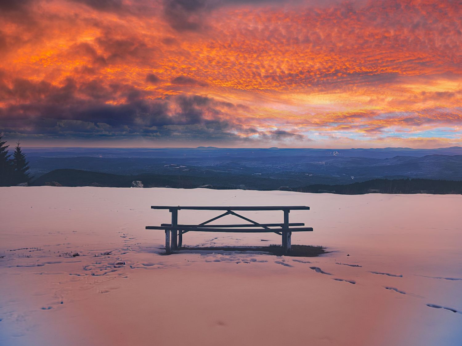 A lone picnic table sits in snow beneath a dramatic fiery orange and red sunset sky over mountain valleys.
