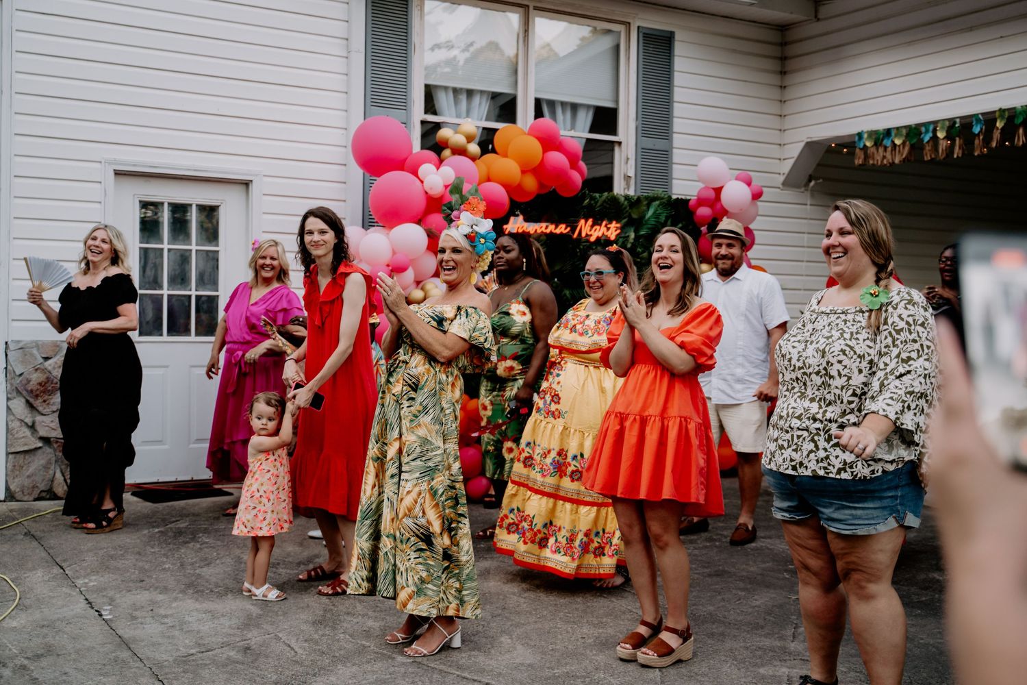A festive group celebrates at an outdoor party with bright pink balloons and colorful floral dresses.