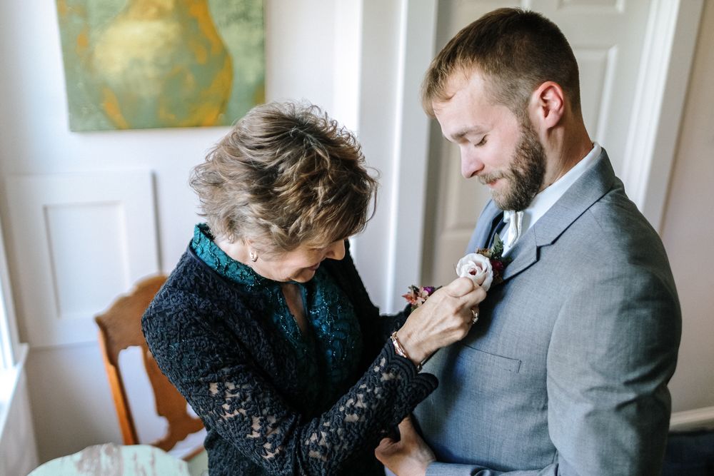 Mother of groom wears black and green sparkling outfit, puts on son's corsage in white walled room with a green and yellow pear painting.