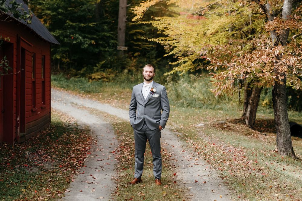 Groom in gray suit stands on dirt gravel path next to red barn with foliage in background. Southern Maine New Hampshire wedding photographer