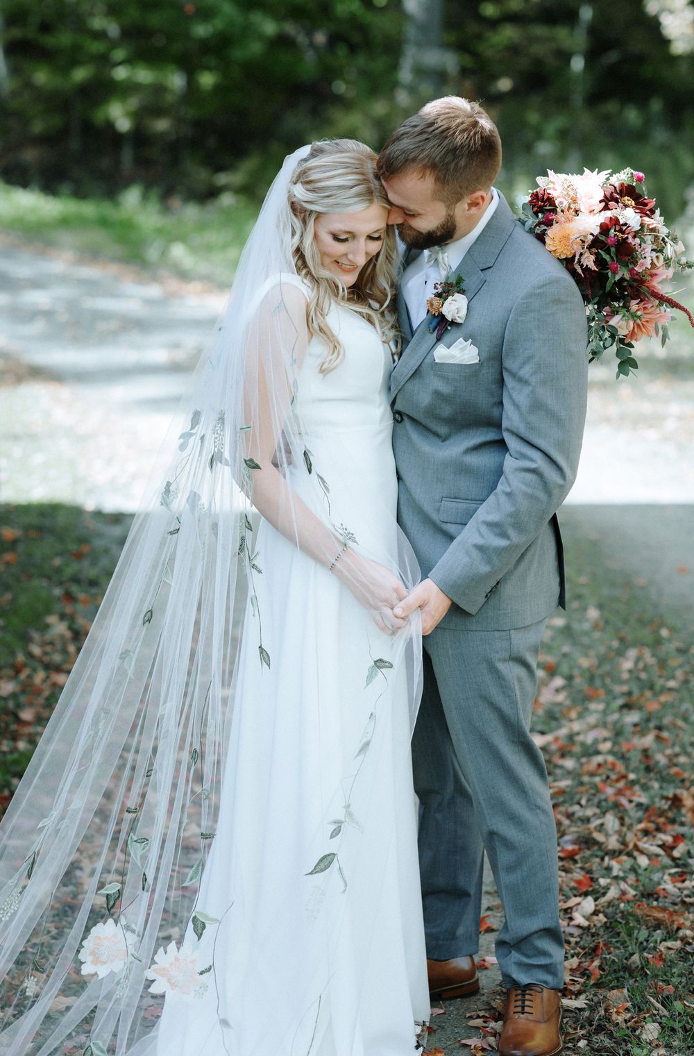 Groom in gray suit embraces bride wearing long veil from side as they smile on their wedding day. Wolfeboro NH wedding photographer.