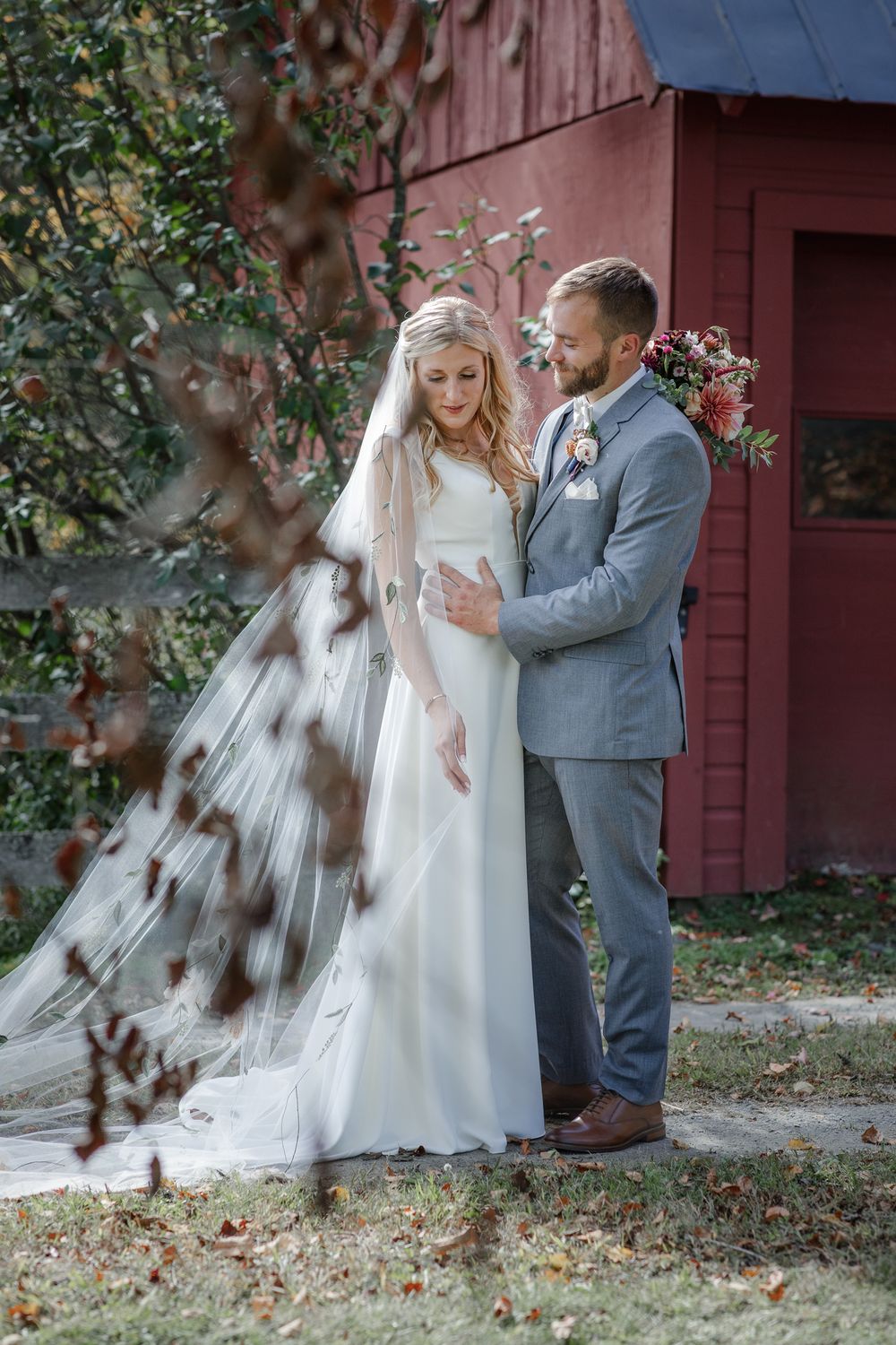 Groom in gray suit embraces bride wearing long veil holding bouquet for First Look photos in front of a red barn at Riverledge Farm Grafton.