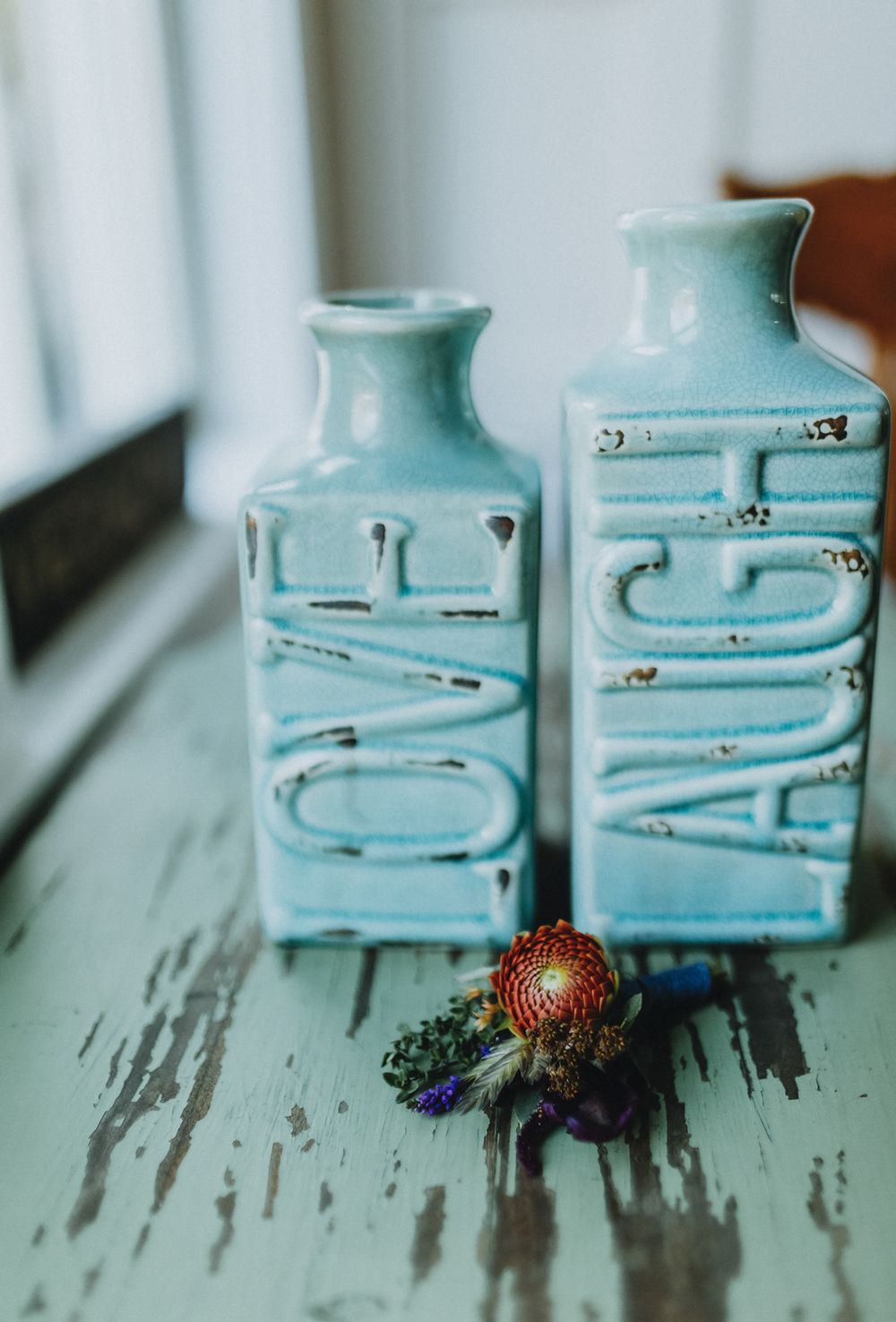 Two light blue ceramic vases with embossed LOVE and LAUGH rests on green wooden table with small boutonniere. Concord wedding photographer.