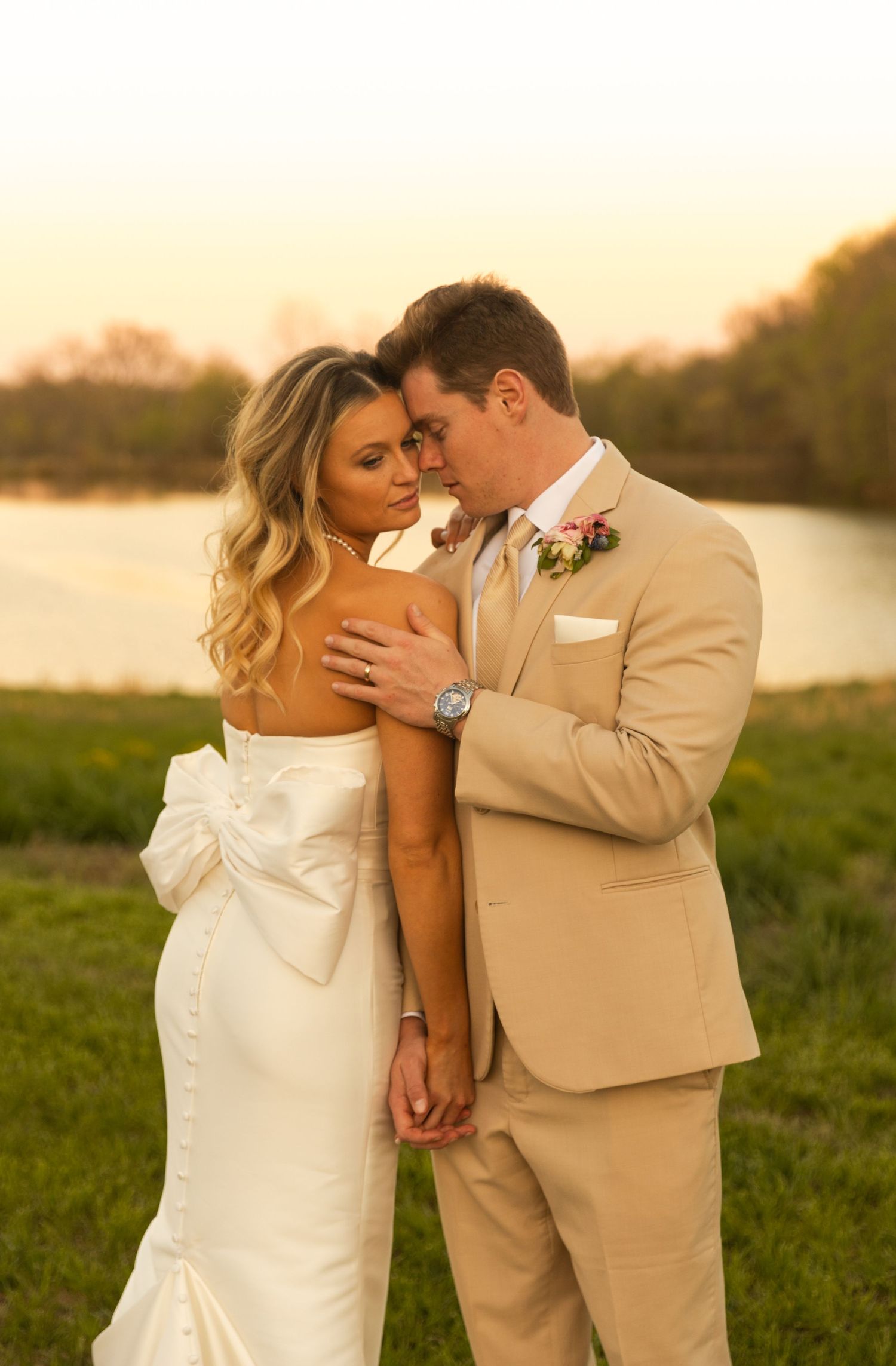 A couple in wedding attire embraces during sunset by a lake.