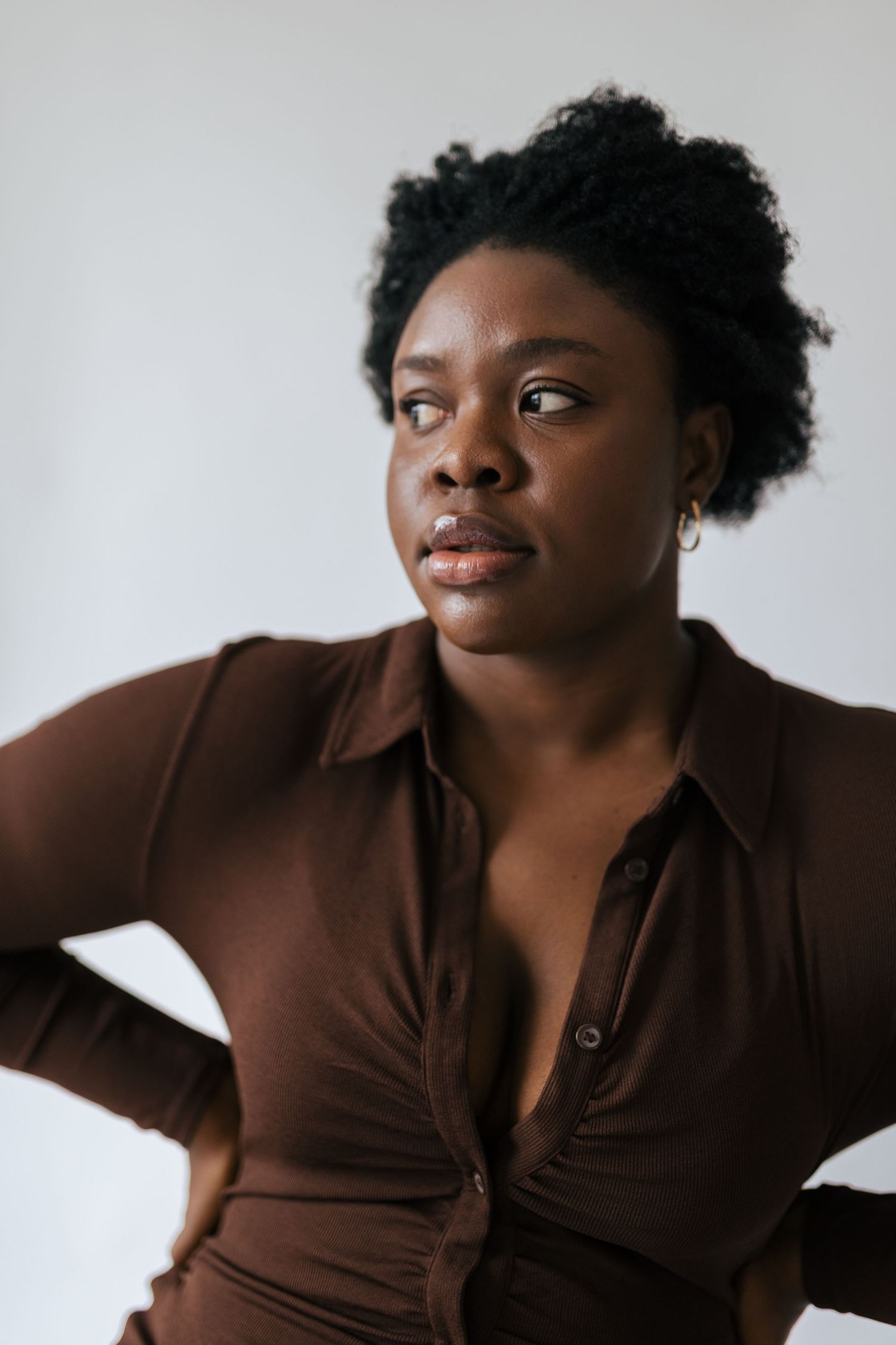 A black woman with natural hair wearing a brown collared top looks out the window in a natural light studio photoshoot.