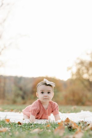 Adorable baby girl in pink knit dress with bow headband crawling on white blanket outdoors in autumn park.