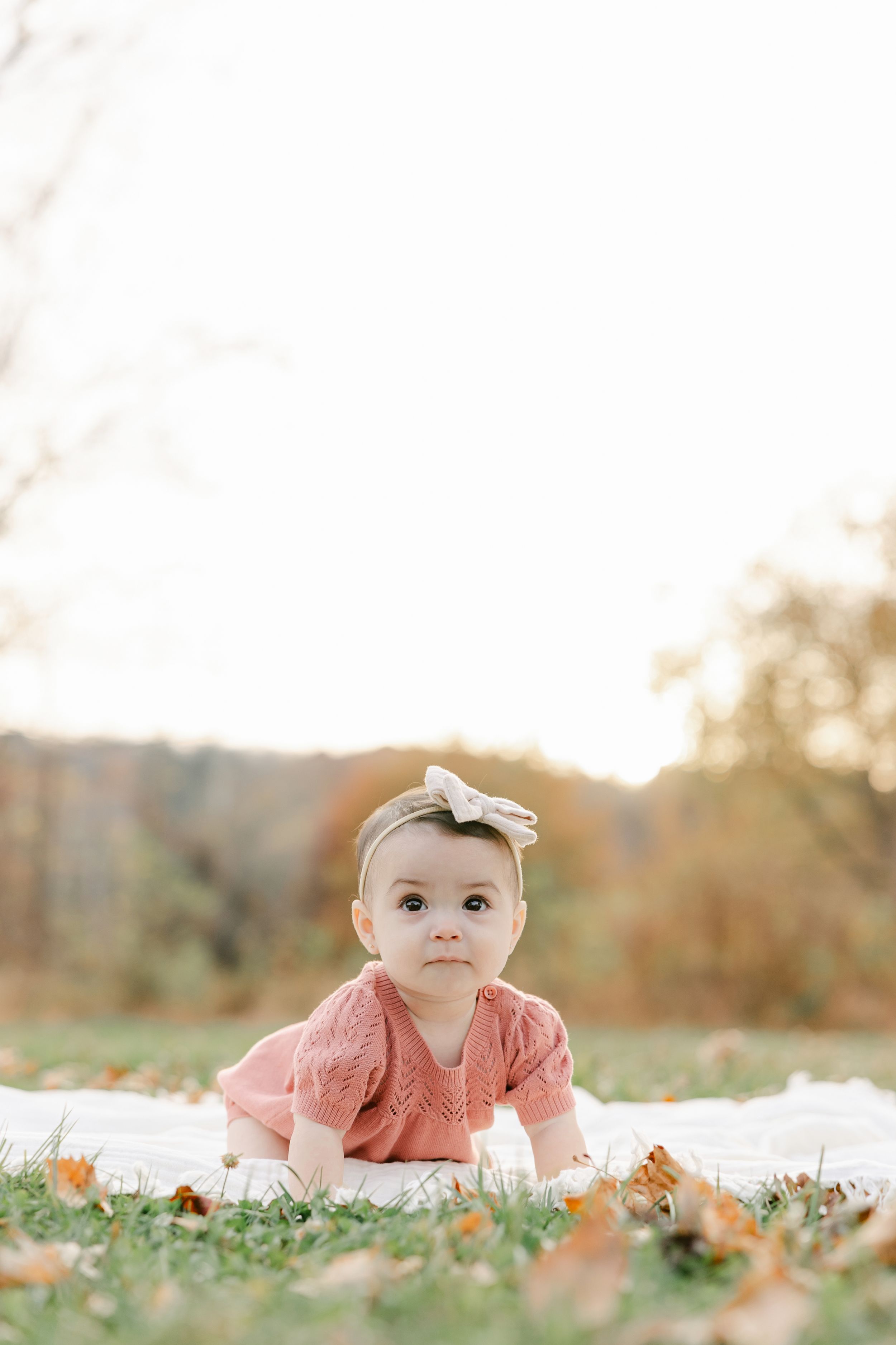 Adorable baby girl in pink knit dress with bow headband crawling on white blanket outdoors in autumn park.