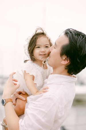 A father kisses his smiling toddler daughter in a white dress, sharing a tender moment outdoors.