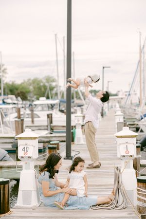 Family enjoying a sunny day on a marina dock, with dad lifting baby while mom and toddler sit together.