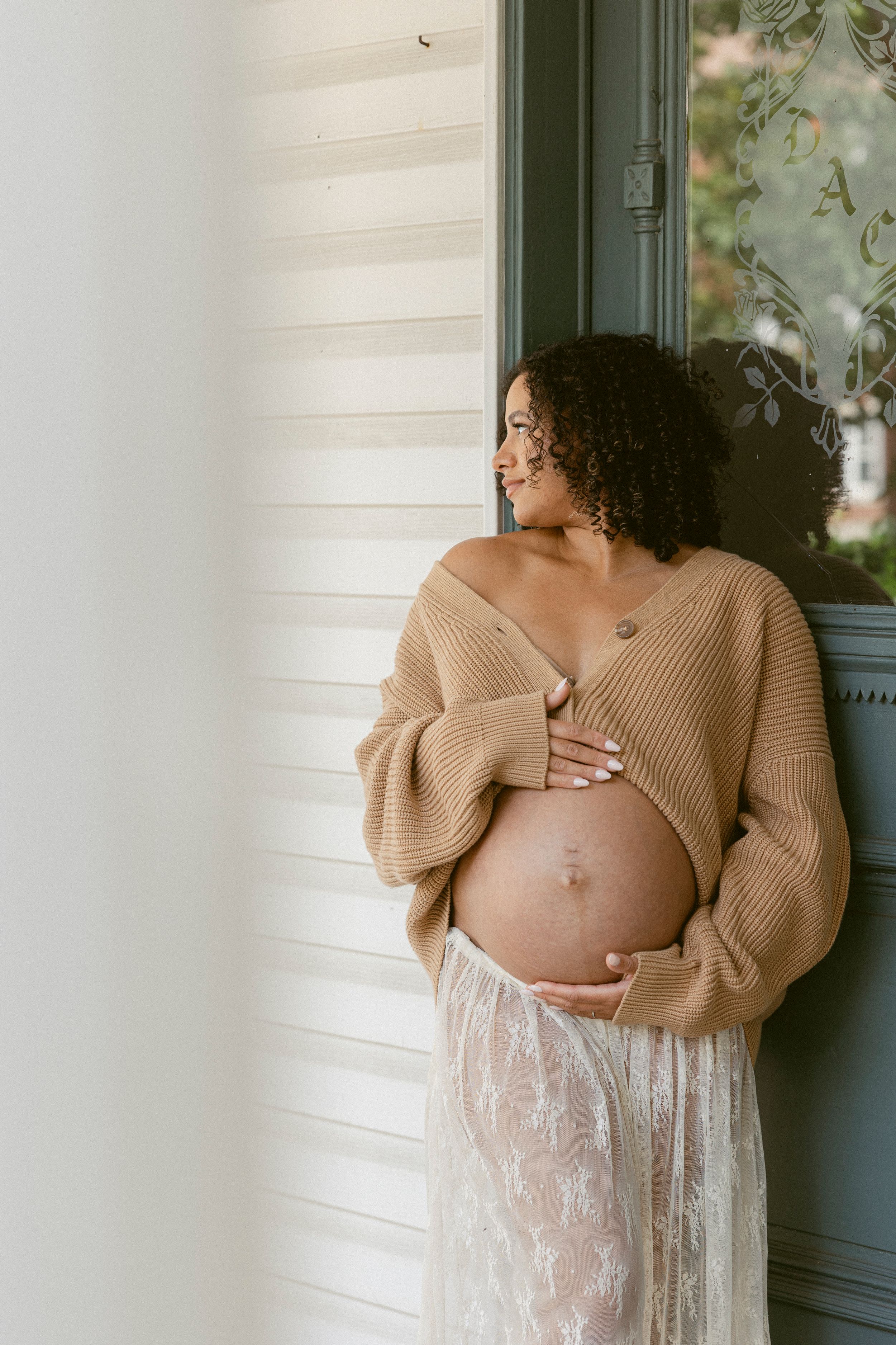 Pregnant woman in camel sweater and lace skirt cradling baby bump while leaning against a door in natural light.