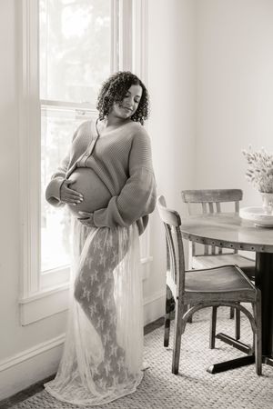 Black and white maternity photo of a pregnant woman in a lace skirt cradling her belly by a bright window.