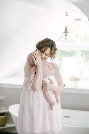 A mother in a pink dress tenderly holds her newborn baby in a bright, airy bathroom setting.
