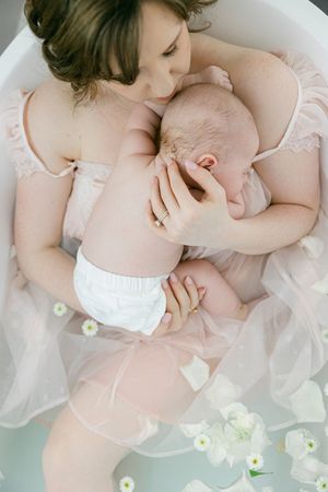Mother in pink dress cradling newborn baby in a floral milk bath, a serene and tender maternity moment.