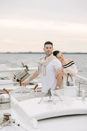 Romantic couple embracing on a yacht deck at sunset, man steering ship wheel, woman in off-shoulder top leaning on his back.