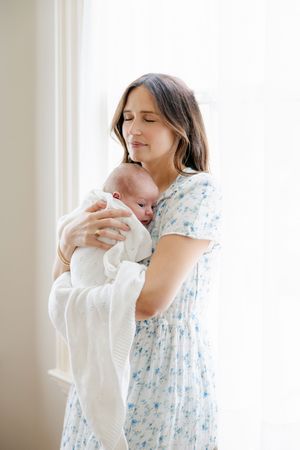 Mother in floral dress holding sleeping newborn by sunlit window. Newborn photography session in CT studio by Ashley Nielsen Studio.