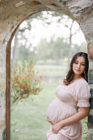 Person in a pink dress poses under a stone archway in a garden setting. Newborn photography session in Connecticut by Ashley Nielsen Studio.