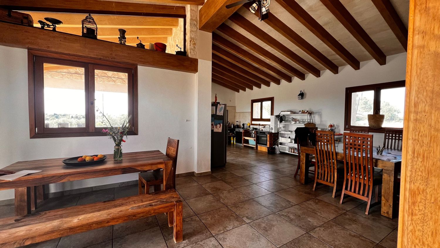 Rustic dining room with wooden furniture, beamed ceiling, tiled floors, and an open kitchen in the background.