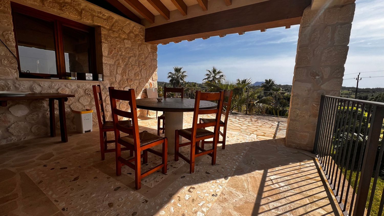 Stone terrace with wooden dining set overlooking lush greenery and sea views under a rustic covered porch.