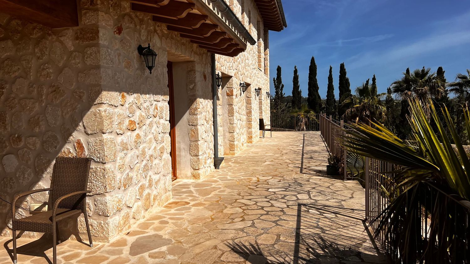 Stone-paved terrace of a Mediterranean villa with rustic walls, cypress trees, and palm plants under a clear blue sky.