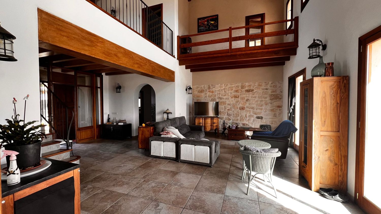 Spacious living room with stone walls, high ceilings, wooden mezzanine balcony, modern furniture, and large tiled floors.