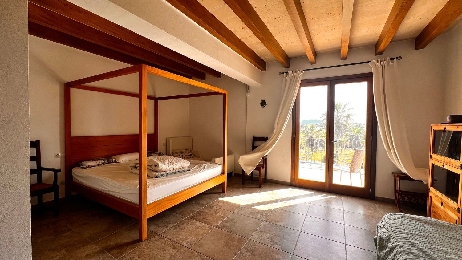 Rustic bedroom with wooden four-poster bed, beamed ceiling, tiled floor, and glass doors opening to a sunny balcony view.