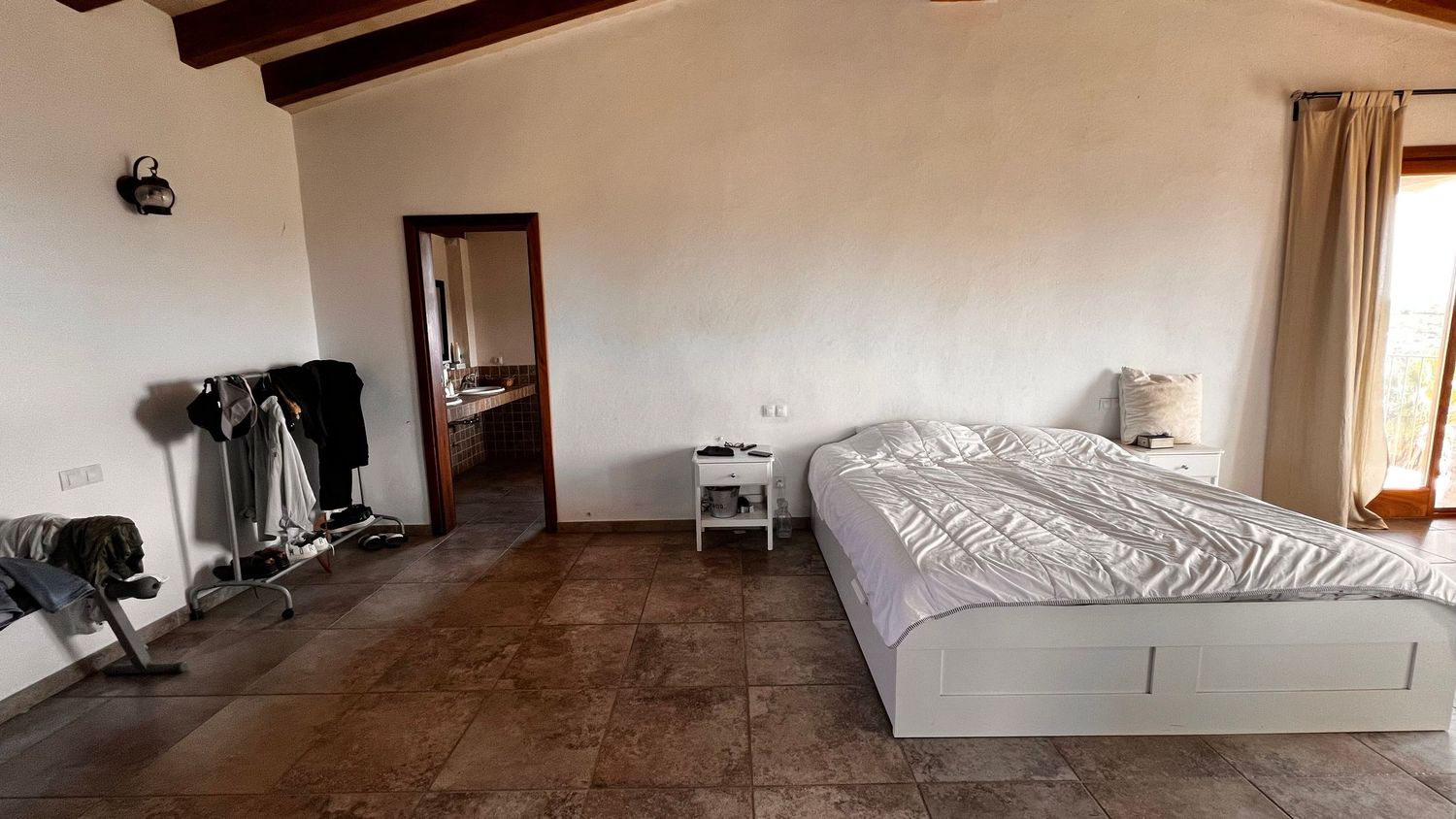 Spacious rustic bedroom with terracotta tile floors, white walls, a large bed, and exercise equipment near an open doorway.