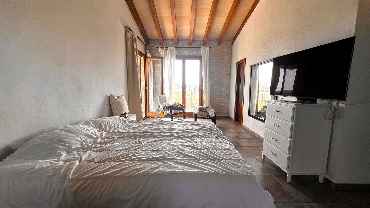 Bright bedroom with wooden beam ceiling, large bed, wall-mounted TV, white dresser, and a window with natural light.